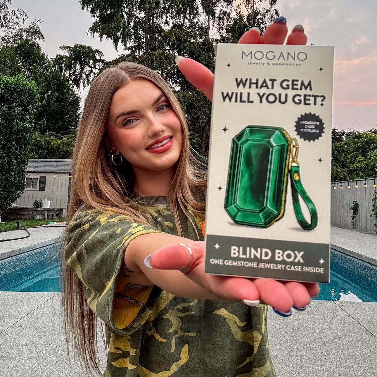 A woman in a green camouflage shirt holds up a 'Mogano Gemstone Jewelry Box' package in front of a swimming pool and trees at sunset.