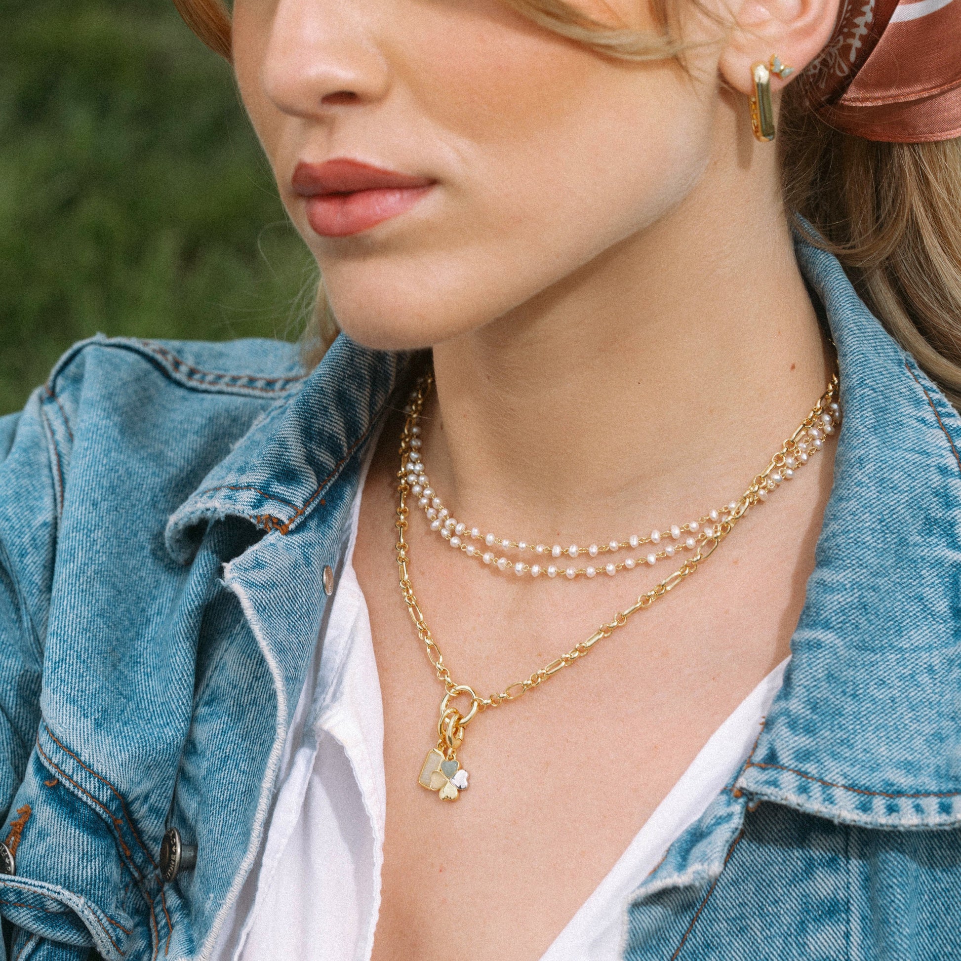 Woman wearing a denim jacket, pearl necklace, and gold earrings with a blurred green background