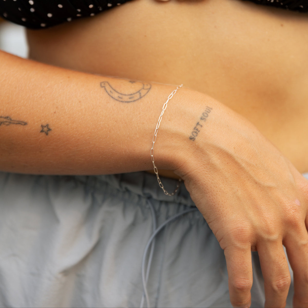 A close-up of a person's torso and arm wearing a delicate silver chain bracelet on the wrist, styled with a black and white polka dot bikini top and light gray drawstring pants.
