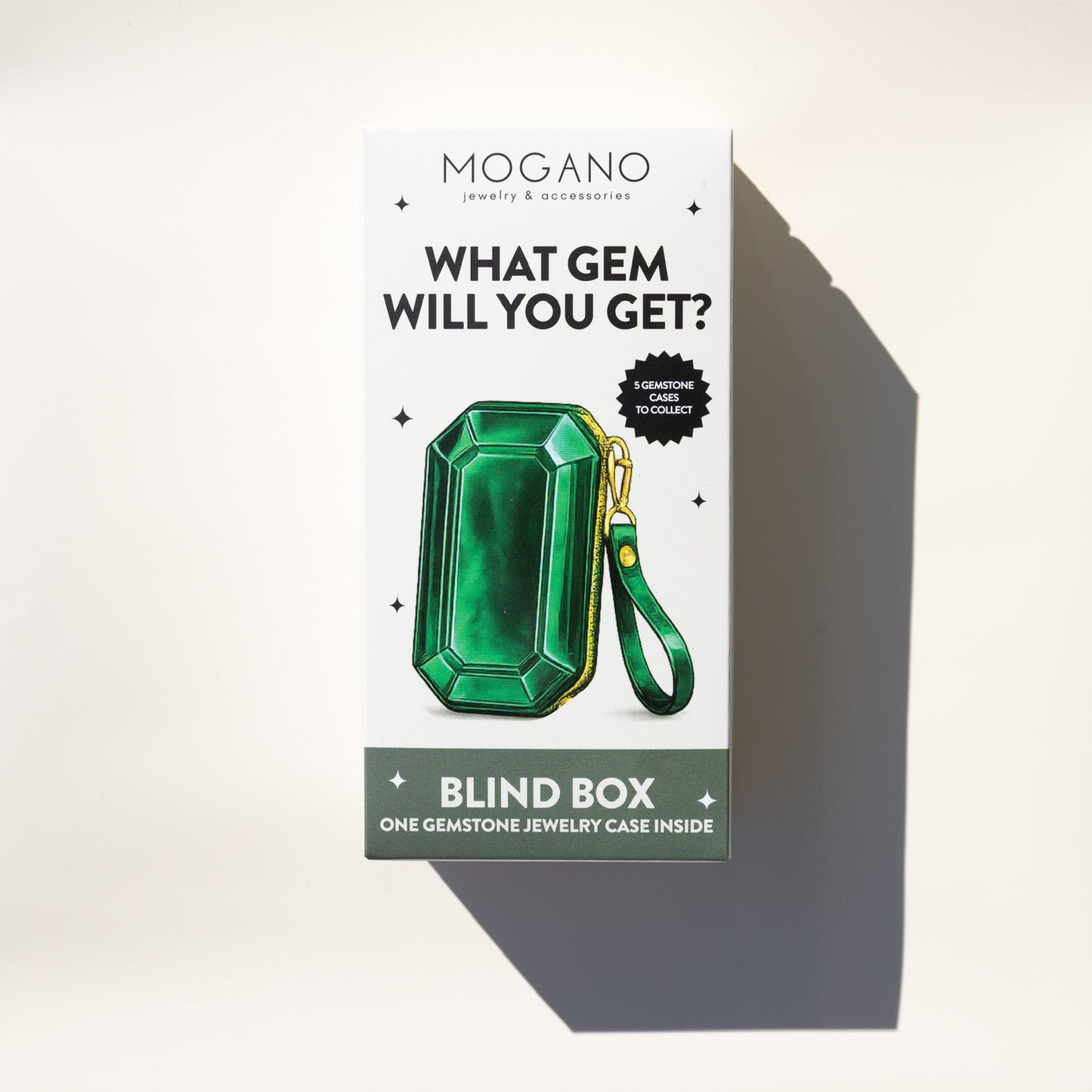 Mogano Jewelry & Accessories 'What Gem Will You Get?' blind box packaging, featuring a green emerald-shaped gem illustration on a white box with a dark green base, photographed on a light beige background with a strong shadow.