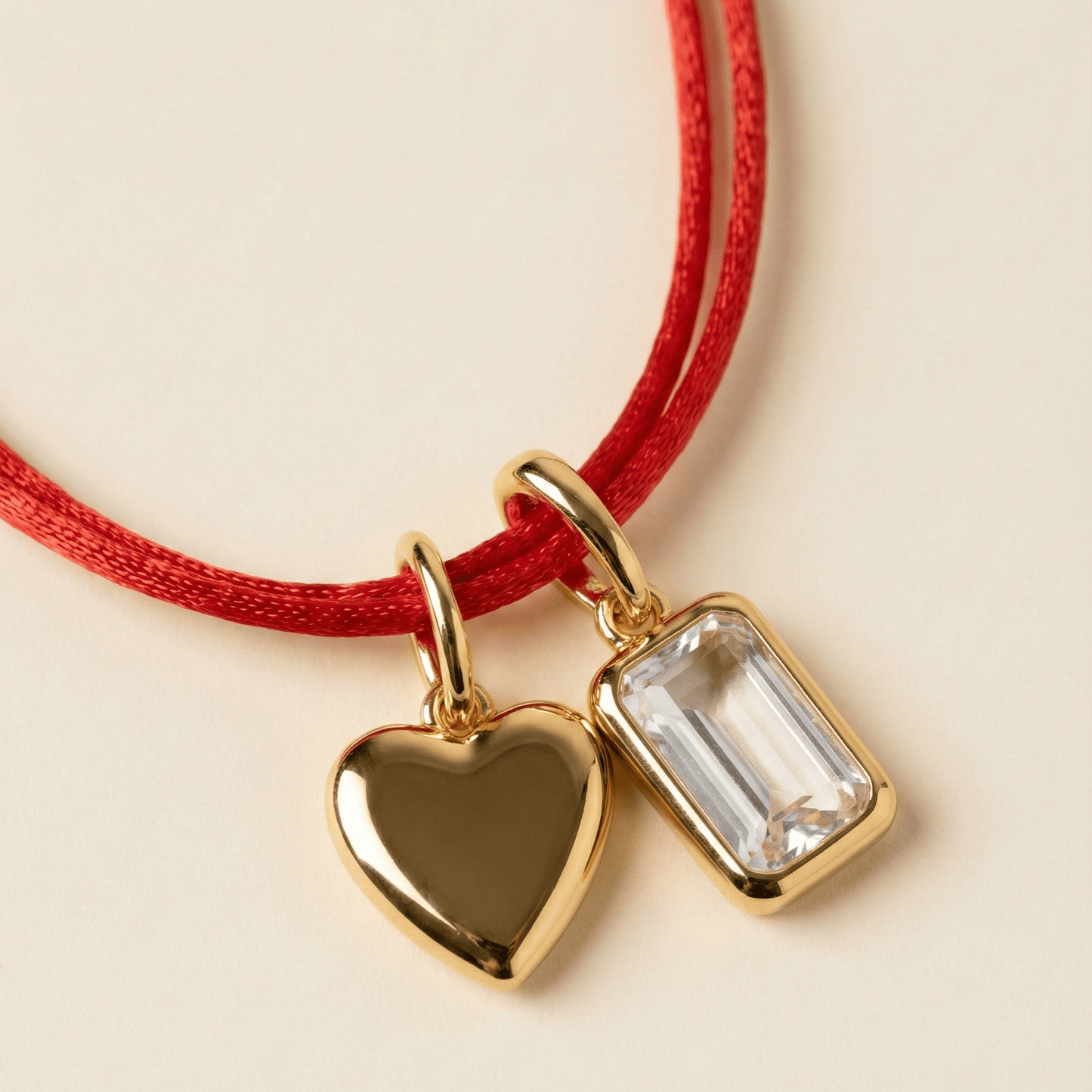 Gold heart and rectangular pendant on a red cord against a beige background