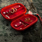 An open red velvet jewelry organizer case with gold-tone rings and accessories inside, placed on a dark rocky surface with water in the background. A red wristlet strap and silver clasp are attached to the case.