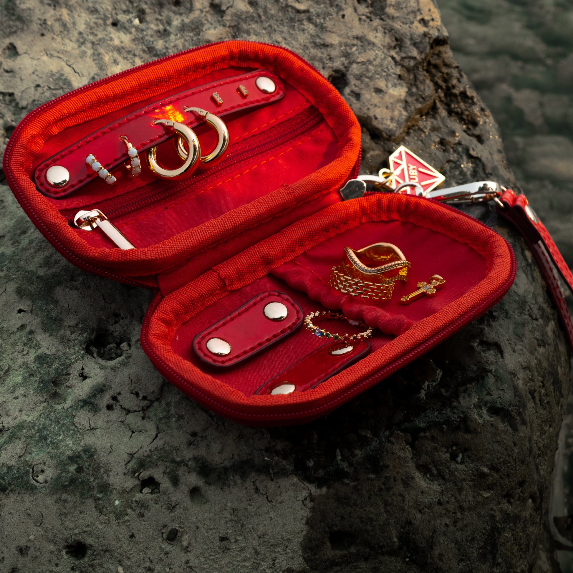 An open red velvet jewelry organizer case with gold-tone rings and accessories inside, placed on a dark rocky surface with water in the background. A red wristlet strap and silver clasp are attached to the case.