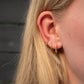 A close-up of a person's ear wearing three small gold hoop earrings, with blonde hair and part of the face visible against a dark gray background.