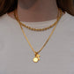 A close-up of a person wearing two layered gold necklaces, including a short curb chain and a longer chain with a small round disc charm and a small cross charm, over a white t-shirt.