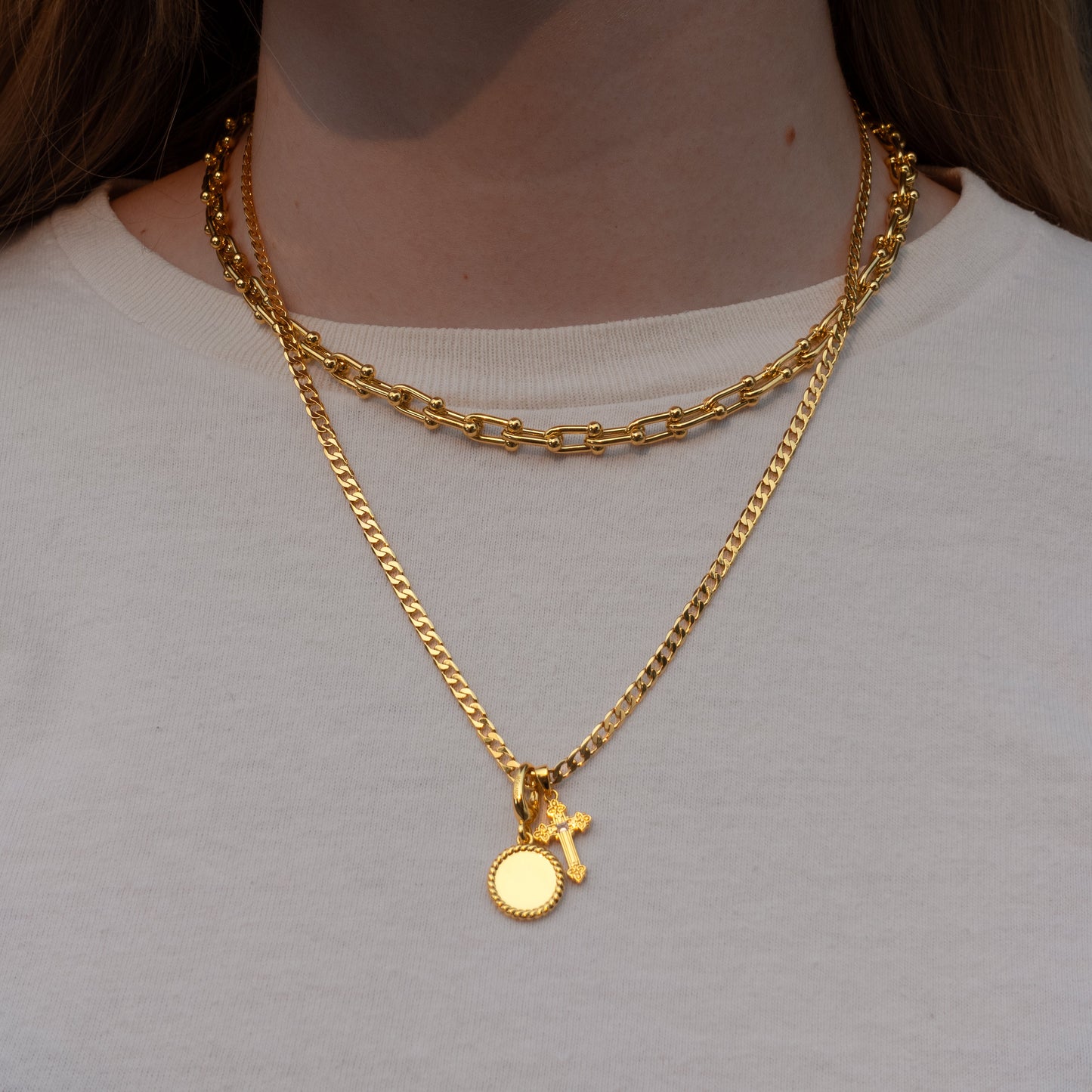 A close-up of a person wearing two layered gold necklaces, including a short curb chain and a longer chain with a small round disc charm and a small cross charm, over a white t-shirt.