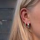 A close-up of a person's ear wearing multiple silver earrings, including a small stud, a cluster of clear stones, and a larger silver hoop, with blonde hair in the foreground.