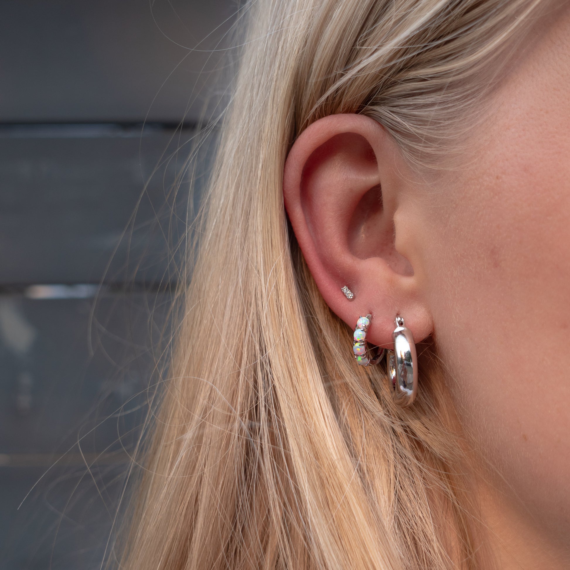 A close-up of a person's ear wearing multiple silver earrings, including a small stud, a cluster of clear stones, and a larger silver hoop, with blonde hair in the foreground.