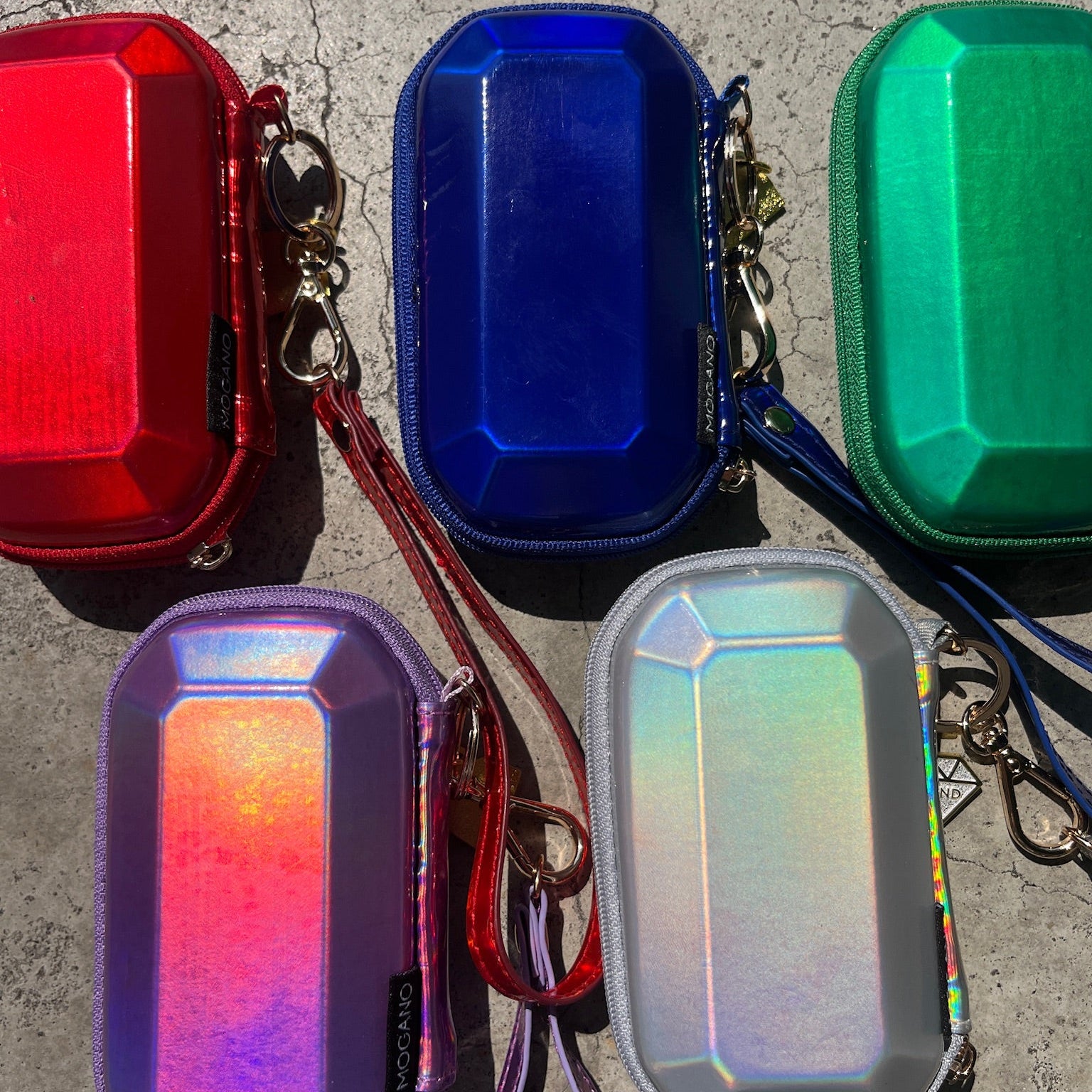 Five small iridescent hard-shell zip pouches or cases arranged on a concrete surface, shown in red, blue, green, purple, and holographic silver with wrist straps and metal clips.