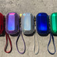 Five small iridescent gem-shaped zip pouches with wrist straps are lined up on a cracked concrete surface. The pouches come in red, purple, silver, blue, and green, with gold-tone hardware.