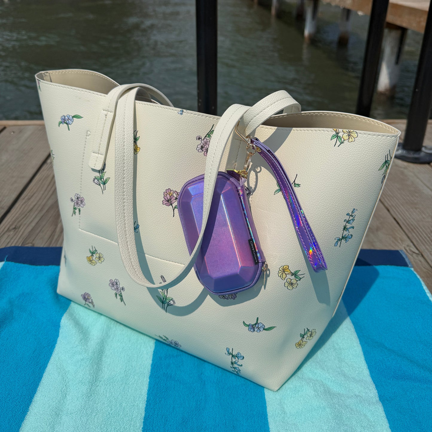 An off-white tote bag with a small floral print, featuring a purple translucent zippered pouch and matching purple zipper pull, resting on a blue and white striped towel next to a canal.