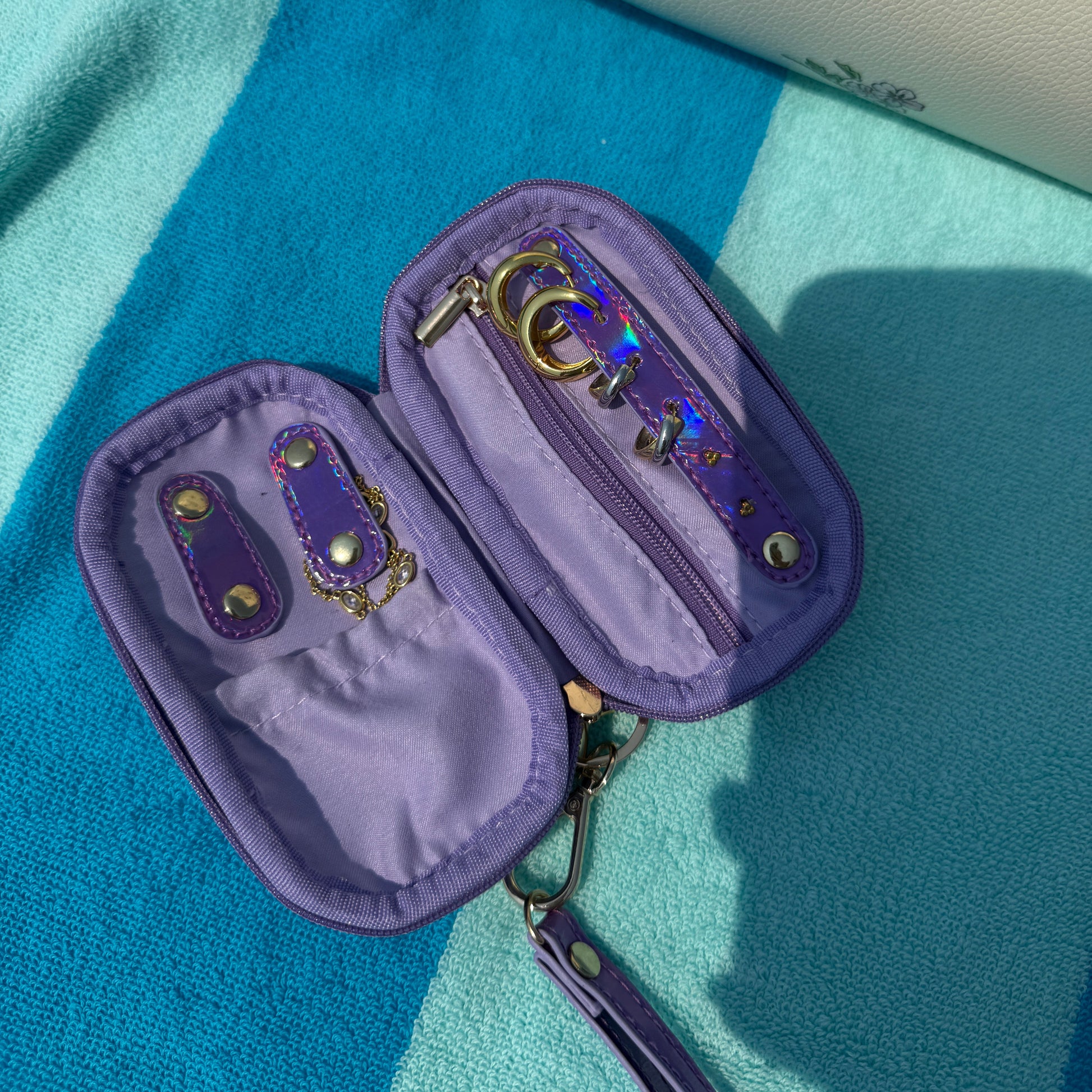 A purple zippered pouch case with gold-tone hardware and iridescent zipper pulls, shown open on a blue and turquoise striped towel with a cream floral bag partially visible in the background.