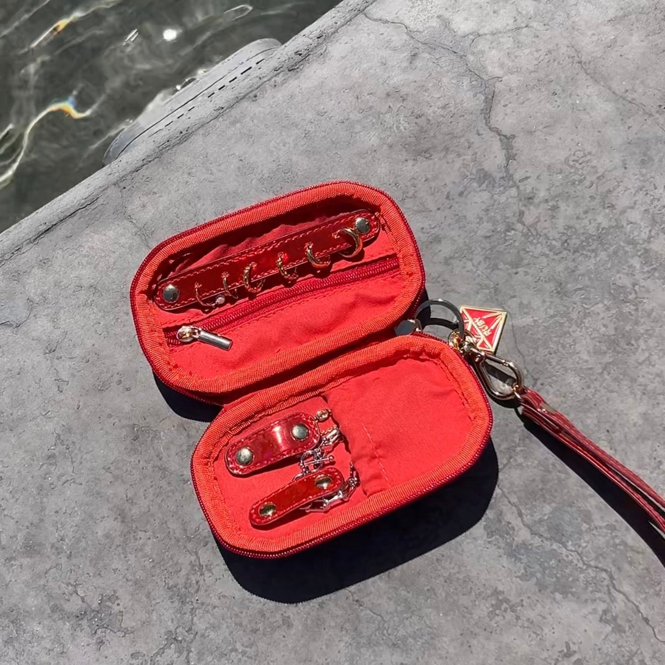 A red nylon and leather travel jewelry case, shown open to display its interior compartments, straps, and metal hardware, jewelry, resting on a gray stone surface next to water.