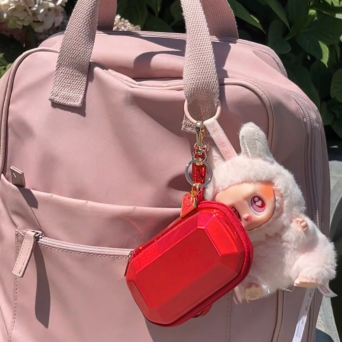 A light pink BEIS canvas backpack with dual top handles, featuring a red zippered pouch jewelry case and a pink plush labubu keychain with pink eyes hanging from the zipper. The backpack is photographed outdoors against a background of green foliage and white flowers.